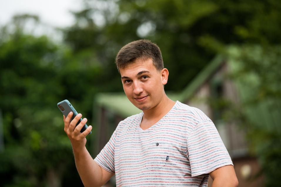 Junger Mann mit kurzen Haaren hält ein Smartphone in der Hand und schaut lächelnd in die Kamera, Hintergrund unscharf.