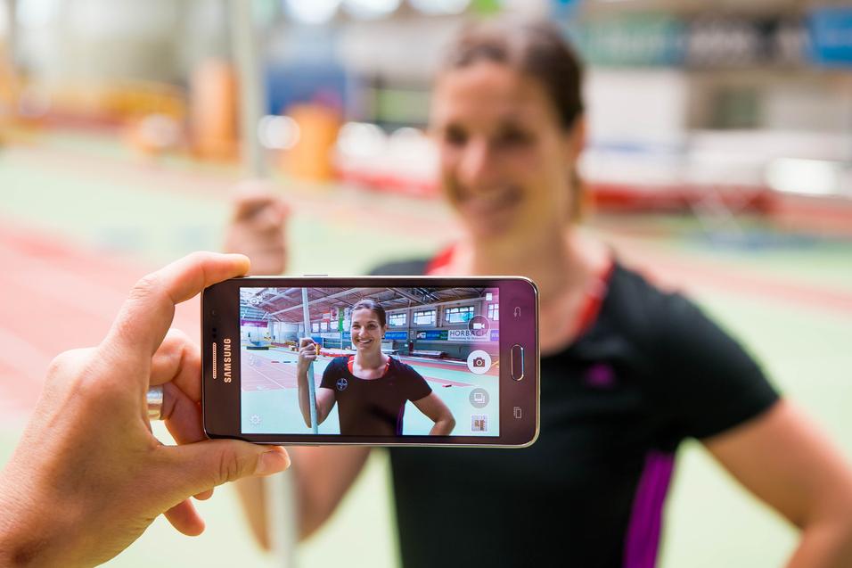Hand hält ein Smartphone, das ein Porträt einer lächelnden Athletin mit Stab in einer Sporthalle zeigt.